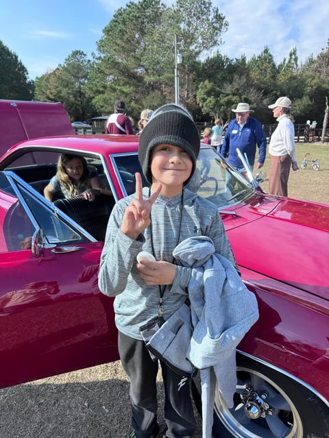 Child in beanie waving at vintage car show with pink 1950s cars and people in background