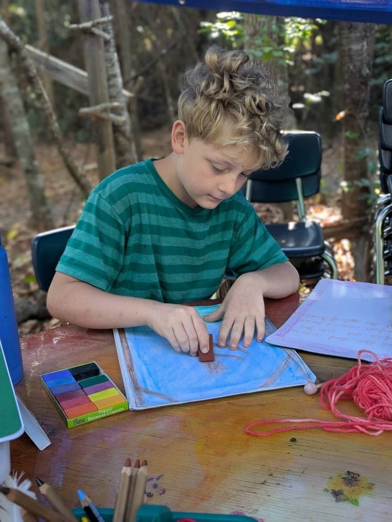 Young child in striped teal shirt painting with watercolors at outdoor table with art supplies