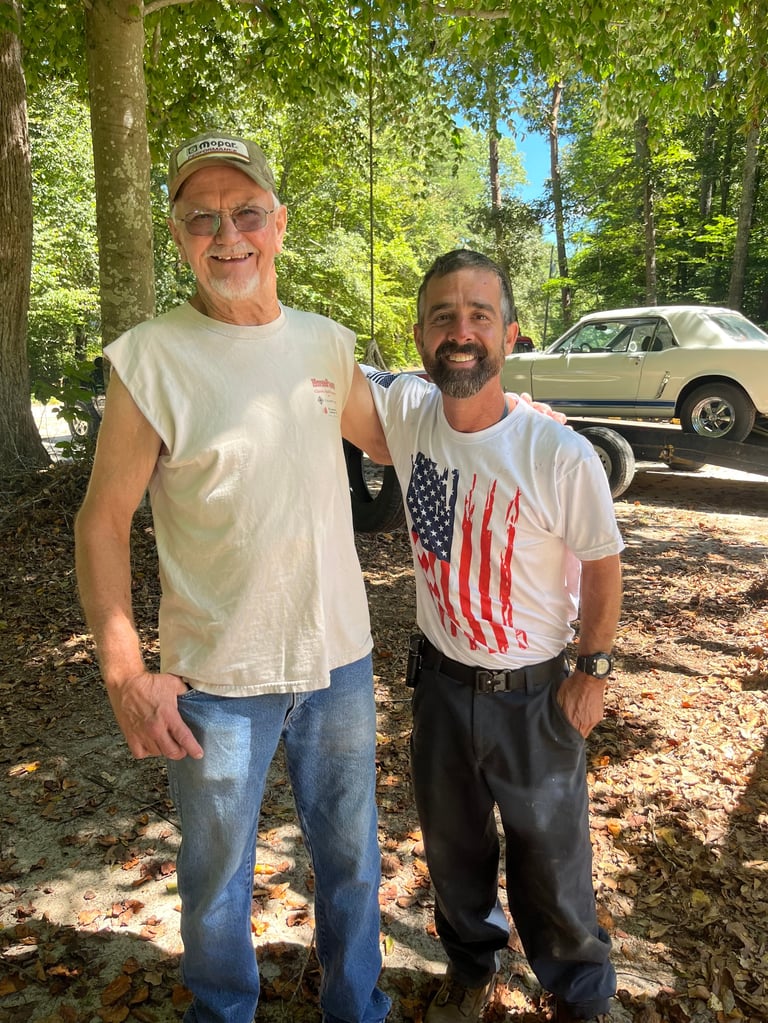 Two men standing outdoors smiling at camera, with vintage cars and trees in background
