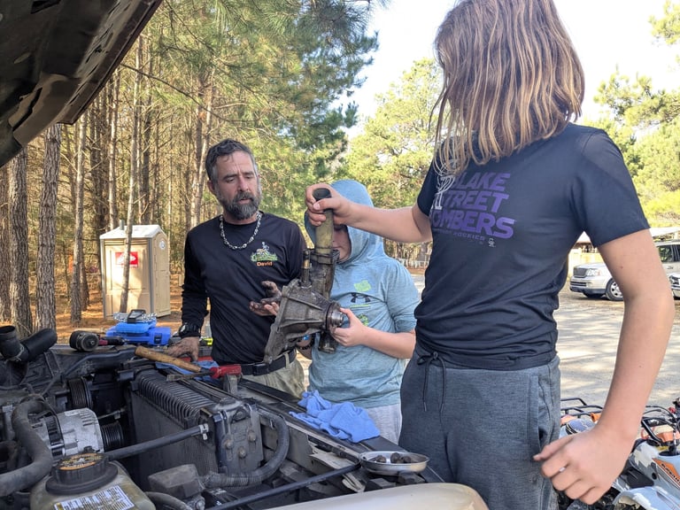 Three people examine an engine component under an open hood at an outdoor automotive workshop