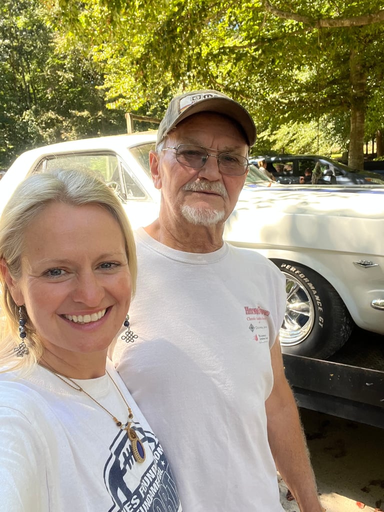 A smiling woman and older man wearing white t-shirts stand together in front of a classic white car and tree-lined background
