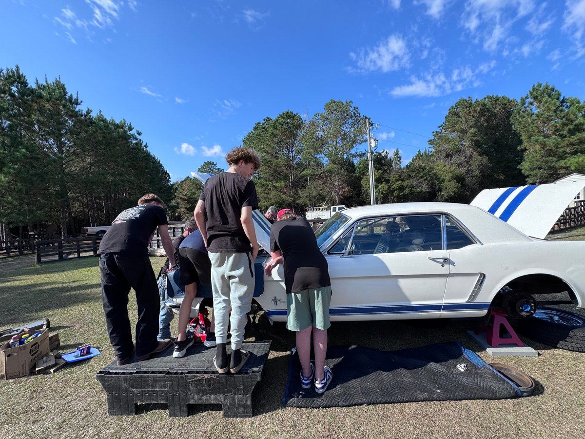 Group of people gathered around a white and blue vintage car at an outdoor car show in a park setting