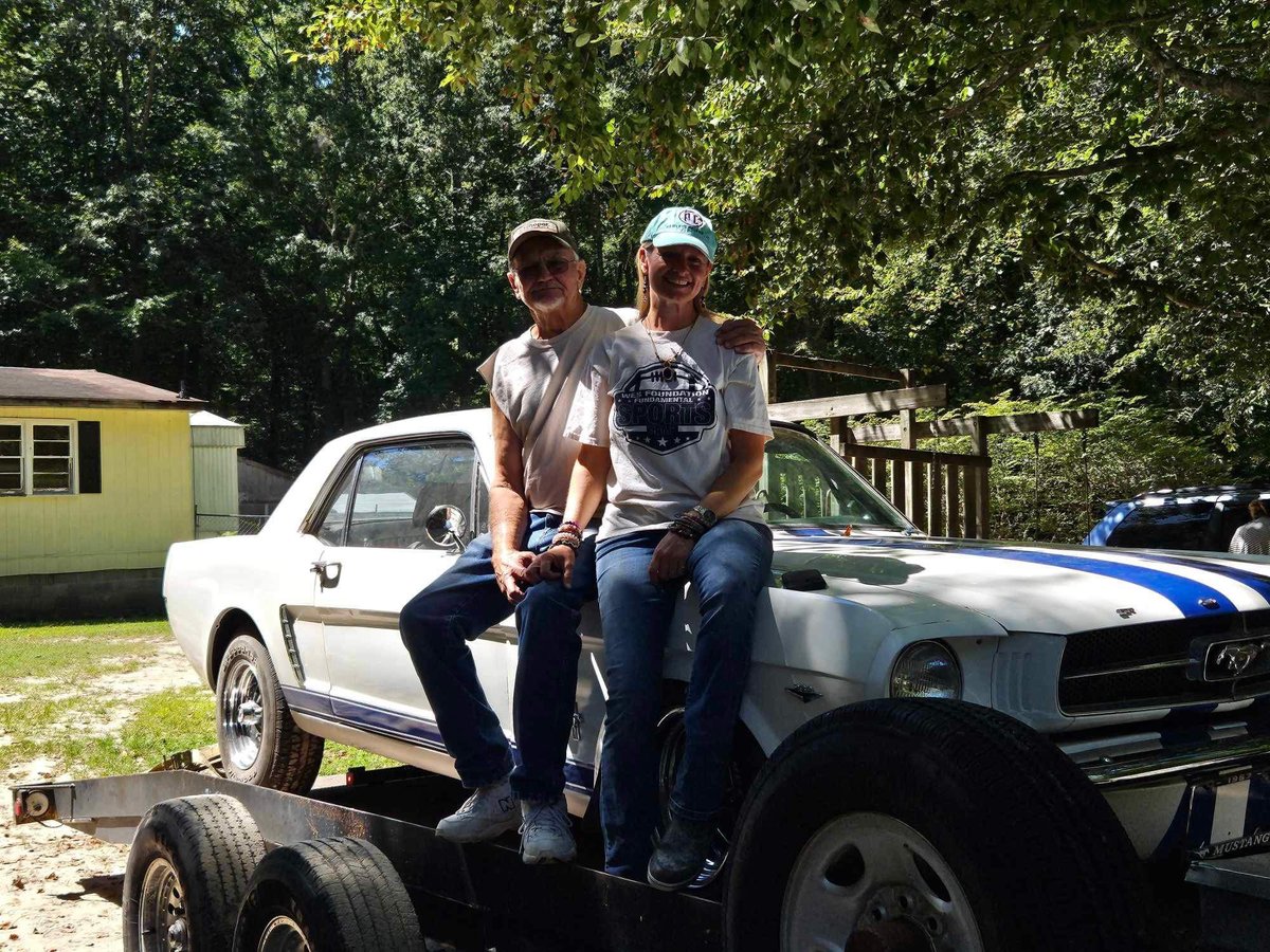 Two men sitting on a classic white Ford Mustang at a car show, with trees and other vehicles in the background