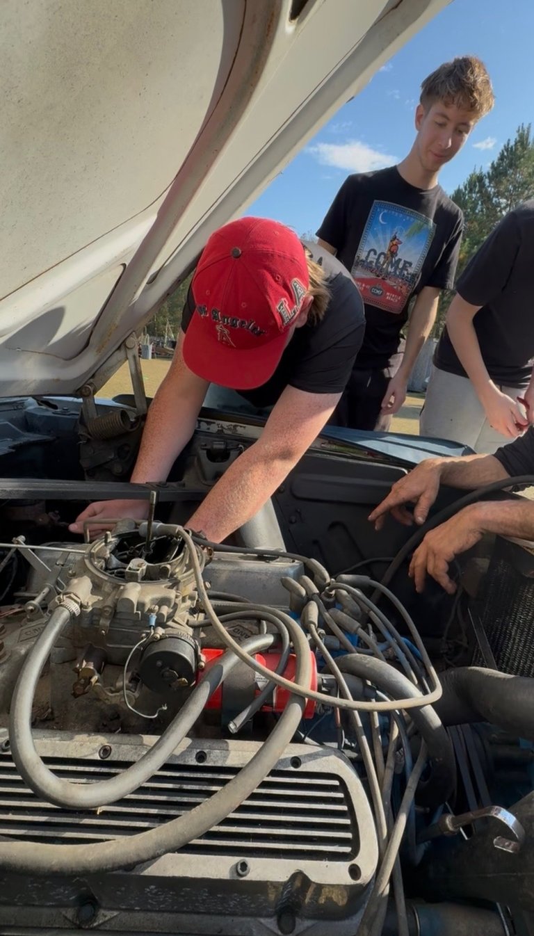Young man in red cap examining car engine at outdoor car show with other people watching