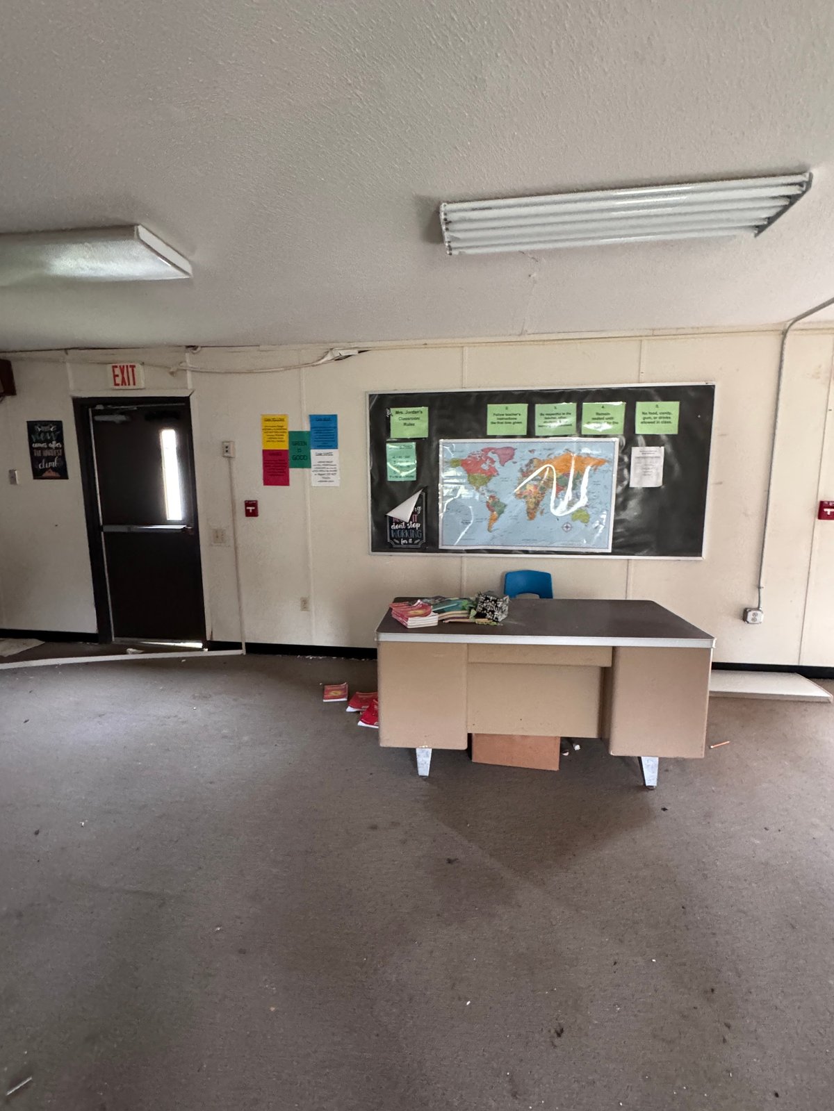 Empty classroom with a central desk, wall-mounted blackboard displaying a world map, and exit door with red emergency sign